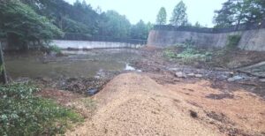 Earthen berm across a shallow river with water pooling behind it; concrete wall on the far side and trees in the background.